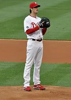 A left-handed man in a white baseball uniform and blue baseball cap with a red brim throws a baseball from a pitcher's mound.
