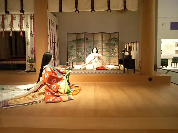 A full-scale model of part of the same palace. The Saiō sits on a tatami dais, with a byōbu behind her, a kichō to her left, and a boxlike chōdai (帳台, baldachin) to her right. Above and before her, a kabeshiro (壁代, wall-curtain) is rolled and tied up. Museum reconstruction of the Heian period's Shinden style.