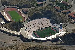 An aerial view of a stadium with blue end zones that appears build into the hills around it. There is a separate track field with seats above and to the left of the stadium.